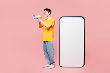 Wall Mural - Full body young man in yellow t-shirt casual clothes big huge blank screen mobile cell phone smartphone with copy space mockup area scream in megaphone isolated on plain pastel light pink background