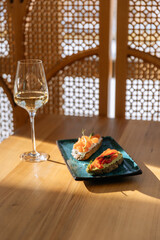 A glass of white wine and red salmon bruschetta in a restaurant on a table in the sun against the background of oriental decor.