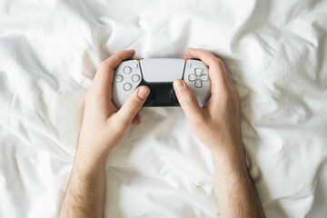 Canvas Print - Man playing video games with game controller while lying in bed on white sheets, top view, gaming console concept.