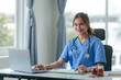 © amnaj - Friendly female nurse in blue scrubs seated at a desk, working on a laptop with medical charts and a stethoscope..