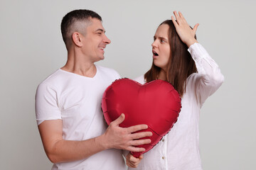 Wall Mural - Smiling man looking at shocked confused woman wearing white clothing holding heart shaped air balloon female forgot about family anniversary isolated over gray background