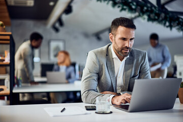 A focused businessman is working over the laptop, with people in the background.