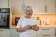 © Shi  - 60s happy woman is using mobile phone standing in the kitchen. High quality photo