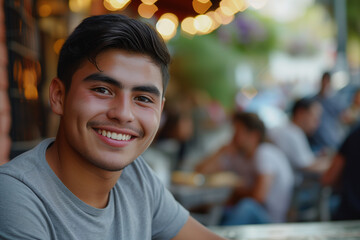 young hispanic man on a restaurant terrace bar smiling teenager latino table lights happy joyful green tshirt plants warm twenties tanned latin brown hair sitting crowd outdoors summer hot smile