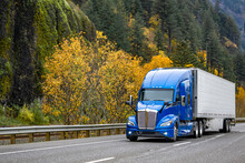 Bonnet blue big rig semi truck tractor carry cargo in refrigerated semi trailer driving on the autumn highway road with rock mountain range on the side