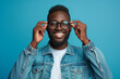 © pprothien - Cheerful African American man adjusting his round glasses, smiling broadly in a denim jacket against a vibrant blue background.