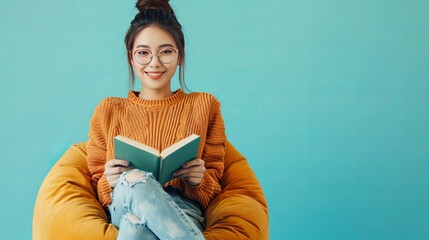 Isolated on a simple pastel light blue-cyan background, a full-body happy Asian woman wears orange sweater spectacles, sits in a bag chair, and reads a book at the library.