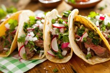 This Is A Photograph Of Four Home Made Steak Tacos With Onions, Radishes, Cilantro, Sauce And Salsa On A Wood Table With A Green And White Checkered Napkin 