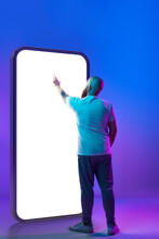 Bald Man In Casual Clothes Pointing At Giant 3D Model Of Mobile Phone Screen Against Gradient Purple Background In Neon Light. Online Services. Concept Of Business, Modern Technology, Gadgets