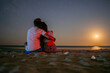A romantic couple embraces from behind on the moonlit sands by the sea, creating an intimate connection under the full moon's luminous glow.