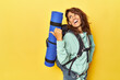 Woman with hiking backpack and mat on yellow points with thumb finger away, laughing and carefree.