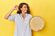 Middle aged woman holding a wall clock on a yellow backdrop feels proud and self confident, example to follow.