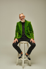 Wall Mural - cheerful bearded attractive mature man in green blazer sitting on tall chair and smiling at camera