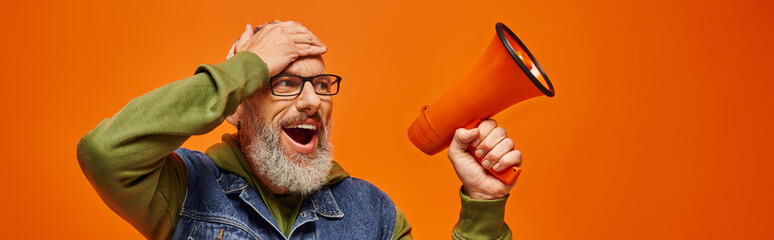 Wall Mural - surprised cheerful mature male model in vibrant fashionable outfit posing with megaphone, banner