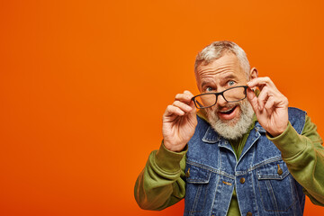 Wall Mural - cheerful bearded mature man in vibrant green hoodie and denim vest posing on orange background