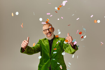 Wall Mural - joyful mature male model in green blazer showing thumbs up under confetti rain on beige background