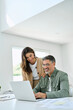 © insta_photos - Happy mature older couple using laptop technology at home. Smiling middle aged senior man working on computer sitting at table with wife standing nearby in living room. Vertical candid shot.