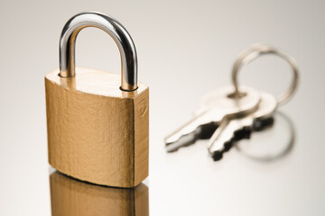 Closed metal padlock in golden color and two keys on a reflective surface. Front view from low angle.