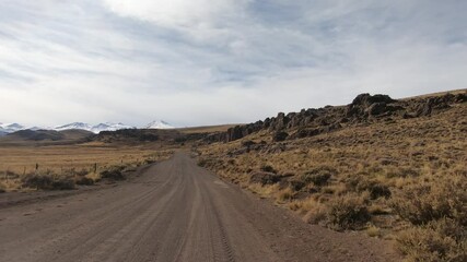 Wall Mural - Traveling in Patagonia. Point of view of a car driving along the dirt road across the yellow grassland and Andes mountains in autumn.