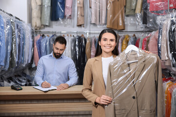 Wall Mural - Dry-cleaning service. Happy woman holding hanger with coat indoors. Worker taking notes at workplace