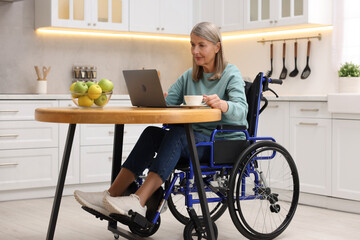 Poster - Woman in wheelchair with cup of drink using laptop at table in home office