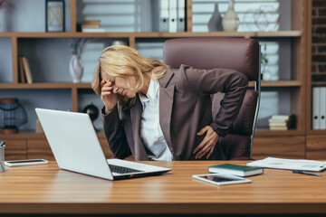 Senior business woman in a suit sitting at a desk in the office holding her hand on her back and head and suffering from severe pain. Needs rest and medical assistance.