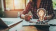 © tasnaifotolia - Conceptual image of a person holding a glowing light bulb with a calculator and paperwork on a desk, symbolizing idea generation and financial planning.