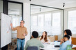 © Vadim Pastuh - In a brightly lit office space, mature man with grey hair leads a discussion in front of a whiteboard, highlighting key points with a marker as his diverse team of young professionals listens intently