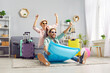 © Studio Romantic - Funny excited happy couple in hats and sunglasses having fun sitting on inflatable mattress ready for holiday trip in living room at home with packed luggage. Vacation and summer journey concept.