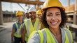 © Tom - Close-up selfie of a cheerful female construction worker in a yellow hard hat and reflective vest, with colleagues in the background on a construction site.
