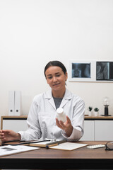 Poster - Female doctor examines bottle of capsules looking with interest young woman in white lab coat smiles recommending medicinal product to patient sitting at table