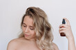 Young caucasian attractive blonde woman with wavy hair spraying thermal protection hairspray, oils with vitamins in a brown bottle on a light background
