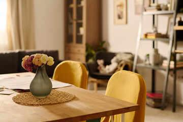 Part of wooden table with grey vase with bunch of flowers surrounded by two yellow chairs against interior of spacious living room