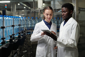 Leinwandbilder - Young engineers of bottled water factory in labcoats looking at tablet screen while discussing information or sketch of machinery