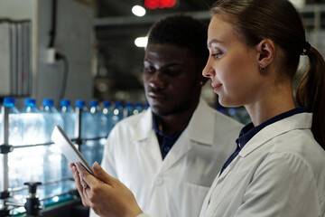 Poster - side view of young female engineer of bottled water factory in labcoat standing next to african amer