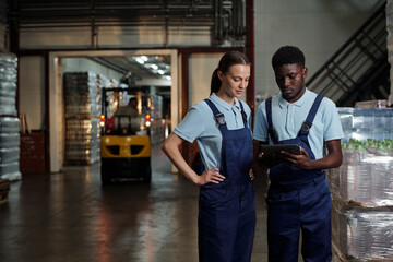 Leinwandbilder - Two young intercultural engineers of quality control looking at screen of tablet while checking or discussing information in warehouse