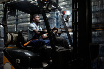 Poster - Young African American man in workwear sitting in forklift and driving it along stacks of packages while holding by steer