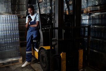 Poster - Young factory engineer in coveralls sitting by door of forklift after work and texting in his smartphone against stacks of bottled water