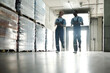 © pressmaster - Blurry outlines of two engineers of bottled water factory moving along stacks of huge packages in spacious distribution warehouse