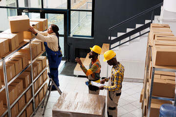 Wall Mural - African american post office employee standing on ladder and taking parcel from shelf. All black package handlers team inspecting order and preparing cardboard box for shipment top view