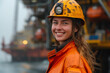 Female engineer. Female offshore engineer wearing a safety helmet and orange protective gear smiles on the deck of an oil rig, with industrial equipment in the background.