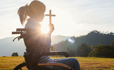 Wall Mural - Christian, Christianity, Religion copy space background. Silhouette girl in wheelchair praying and holding christian cross for worshipping God at sunset background.