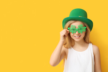 Canvas Print - Cute little girl in leprechaun hat and decorative glasses in shape of clover on yellow background. St. Patrick's Day celebration