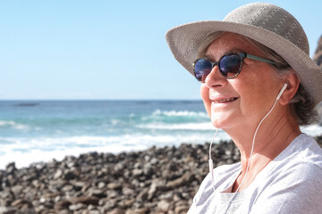 Sticker - Smiling relaxed senior pensioner woman with hat sitting in outdoors at the beach listening music by earphones, elderly female enjoying free time and retirement