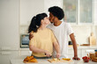 © DragonImages - Two young intercultural sweethearts kissing one another while affectionate woman chopping fresh banana on board by kitchen table