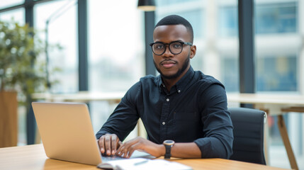 Confident individual seated at a table with a laptop in an office with a city view.