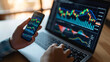 © MP Studio - a person is holding a smartphone with stock market data on the screen, with a blurred background showing a computer monitor with financial graphs