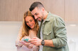 © Carla - Young adult couple marriage using their smartphones at home sharing and smiling watching social media content. Boyfriend and girlfriend texting and planning with the cellphones in their kitchen