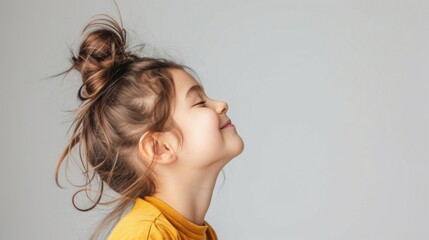 Canvas Print - A young girl with a messy bun smiling and looking up wearing a yellow top against a neutral background.