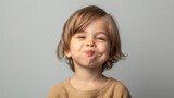 Fototapeta  - A young child with a playful expression sticking out their tongue and smiling against a neutral gray background.